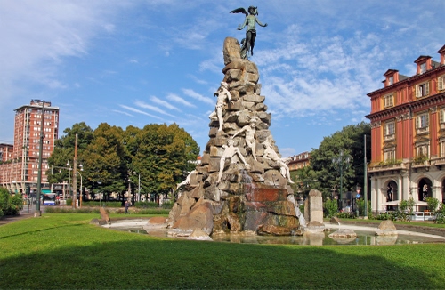 Immagine con alberi, monumento, nuvole, cielo, palazzi, statue, aiuola, verde, fontana, piazza, azzurro, prato, angelo, ali, statua, finestre, giardino, acqua, case, scultura
